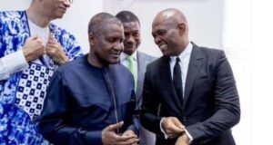 No Rift With Tony Elumelu, Dangote Group Claims Four men in formal clothing chat and smile at an indoor event; two are foreground, one in a navy traditional outfit and one in a black suit.