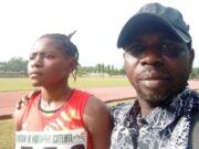 Oyo Athletics Boss Dr. Ogada Narrates The Trajectory Of Taofikat, MTN Champs Athletics’ 200m Winner Two people on a running track posing for a photo; a woman in a red sleeveless top with a race bib stands beside a man wearing a black cap and patterned shirt.