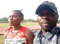 Oyo Athletics Boss Dr. Ogada Narrates The Trajectory Of Taofikat, MTN Champs Athletics’ 200m Winner Two people on a running track posing for a photo; a woman in a red sleeveless top with a race bib stands beside a man wearing a black cap and patterned shirt.