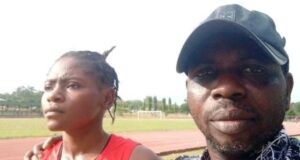 Oyo Athletics Boss Dr. Ogada Narrates The Trajectory Of Taofikat, MTN Champs Athletics’ 200m Winner Two people on a running track posing for a photo; a woman in a red sleeveless top with a race bib stands beside a man wearing a black cap and patterned shirt.