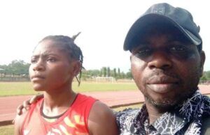 Oyo Athletics Boss Dr. Ogada Narrates The Trajectory Of Taofikat, MTN Champs Athletics’ 200m Winner Two people on a running track posing for a photo; a woman in a red sleeveless top with a race bib stands beside a man wearing a black cap and patterned shirt.