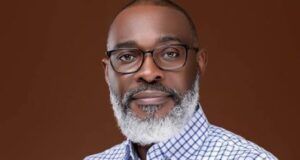 Tinubu Appoints Retired Army General As Special Adviser On Homeland Security Portrait of a mature man with glasses and a gray beard, wearing a blue checkered shirt against a solid brown background.