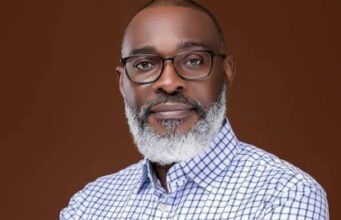 Tinubu Appoints Retired Army General As Special Adviser On Homeland Security Portrait of a mature man with glasses and a gray beard, wearing a blue checkered shirt against a solid brown background.