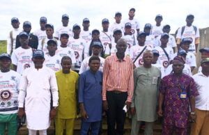 TECH-U VC Commends Hon. Oluwafowokanmi At Flag-Off Of Technical Trainers Recruitments, Training Programme Group of graduates in white T-shirts and caps posing on steps outdoors, with a few officials in colorful outfits standing at the front.