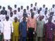 TECH-U VC Commends Hon. Oluwafowokanmi At Flag-Off Of Technical Trainers Recruitments, Training Programme Group of graduates in white T-shirts and caps posing on steps outdoors, with a few officials in colorful outfits standing at the front.