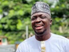 APC Primaries: Abdulganeey Adekunle, ‘Mr. GRA,’Secures House Of Assembly Ticket Smiling man wearing a white traditional outfit with gold trim and a patterned cap outdoors in greenery.
