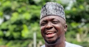 APC Primaries: Abdulganeey Adekunle, ‘Mr. GRA,’Secures House Of Assembly Ticket Smiling man wearing a white traditional outfit with gold trim and a patterned cap outdoors in greenery.