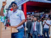 Gov. Makinde Is Capable Of Leading Nigeria — Oyo ALGON Chairman Man at a podium raising his finger while speaking at an outdoor rally, with a crowd and officials behind him.