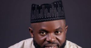 APC House Of Assembly Hopeful, Giwa, Salutes Workers On May Day Portrait of a bearded man in a light beige traditional outfit and a black patterned hat, with his hands clasped under his chin and looking at the camera.