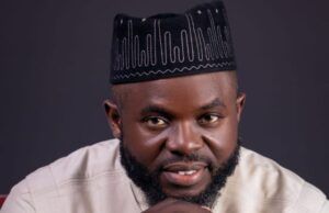 APC House Of Assembly Hopeful, Giwa, Salutes Workers On May Day Portrait of a bearded man in a light beige traditional outfit and a black patterned hat, with his hands clasped under his chin and looking at the camera.