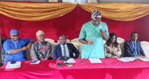 OYSG Trains 35 Workers In Human Capacity Building Panel of six people seated at a red table on a stage with gold drapery; a man in a green shirt speaks into a microphone while others listen and prepare notes.