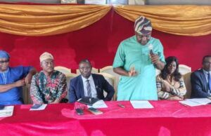 OYSG Trains 35 Workers In Human Capacity Building Panel of six people seated at a red table on a stage with gold drapery; a man in a green shirt speaks into a microphone while others listen and prepare notes.