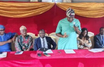 OYSG Trains 35 Workers In Human Capacity Building Panel of six people seated at a red table on a stage with gold drapery; a man in a green shirt speaks into a microphone while others listen and prepare notes.