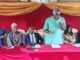 OYSG Trains 35 Workers In Human Capacity Building Panel of six people seated at a red table on a stage with gold drapery; a man in a green shirt speaks into a microphone while others listen and prepare notes.