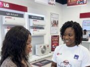 LG Electronics Expands Health Protection For Nigerian Consumers With Free Comprehensive Insurance Two women in an LG store; a sales associate wearing a 'Free Malaria Insurance' shirt shows a tablet to a customer, with LG air-conditioners on the wall behind them.