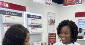 LG Electronics Expands Health Protection For Nigerian Consumers With Free Comprehensive Insurance Two women in an LG store; a sales associate wearing a 'Free Malaria Insurance' shirt shows a tablet to a customer, with LG air-conditioners on the wall behind them.