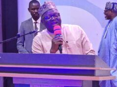 NOA DG Advocates Positive Mindset, Responsible Communication For National Development At UNIFEMGA Reunion Man in traditional cap speaking into a pink microphone at a lectern on a stage, with two men in the background.
