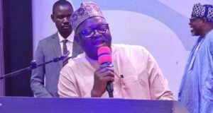 NOA DG Advocates Positive Mindset, Responsible Communication For National Development At UNIFEMGA Reunion Man in traditional cap speaking into a pink microphone at a lectern on a stage, with two men in the background.