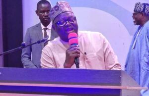 NOA DG Advocates Positive Mindset, Responsible Communication For National Development At UNIFEMGA Reunion Man in traditional cap speaking into a pink microphone at a lectern on a stage, with two men in the background.