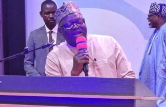 NOA DG Advocates Positive Mindset, Responsible Communication For National Development At UNIFEMGA Reunion Man in traditional cap speaking into a pink microphone at a lectern on a stage, with two men in the background.