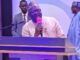 NOA DG Advocates Positive Mindset, Responsible Communication For National Development At UNIFEMGA Reunion Man in traditional cap speaking into a pink microphone at a lectern on a stage, with two men in the background.