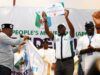 Gov. Makinde Emerges APM Consensus Presidential Candidate Man in white shirt and cap raises a certificate on a stage as others applaud, a banner reading 'People's Movement' behind them indicating an awards ceremony or recognition event.