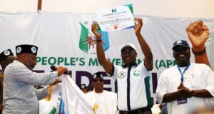 Gov. Makinde Emerges APM Consensus Presidential Candidate Man in white shirt and cap raises a certificate on a stage as others applaud, a banner reading 'People's Movement' behind them indicating an awards ceremony or recognition event.