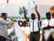 2027: Makinde’s Growing Popularity Signals Presidential Victory For APM—Party Man in white shirt and cap raises a certificate on a stage as others applaud, a banner reading 'People's Movement' behind them indicating an awards ceremony or recognition event.