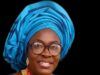 Mama Empowerment Foundation Plans Mega July Programme In Ibadan Smiling woman in a bright blue head wrap with glasses, gold earrings, and a red beaded necklace against a dark background.