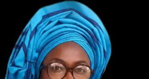 Mama Empowerment Foundation Plans Mega July Programme In Ibadan Smiling woman in a bright blue head wrap with glasses, gold earrings, and a red beaded necklace against a dark background.
