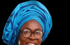 Mama Empowerment Foundation Plans Mega July Programme In Ibadan Smiling woman in a bright blue head wrap with glasses, gold earrings, and a red beaded necklace against a dark background.