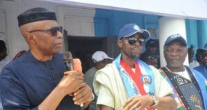 Tinubu’s Performance Influenced My Defection To APC—Mimiko Three men at a public event: the leftmost man in a black cap and dark clothing speaks into a microphone, while two others in colorful outfits and caps stand beside him.