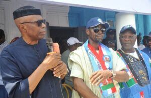 Tinubu’s Performance Influenced My Defection To APC—Mimiko Three men at a public event: the leftmost man in a black cap and dark clothing speaks into a microphone, while two others in colorful outfits and caps stand beside him.