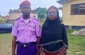 NGO Partners Oyo NOA On Civic Re-Engineering, Responsible Citizenship Man in a purple security uniform with a red beret stands next to a woman wearing a black hijab outdoors near buildings and grass.