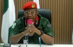 Oyo NSCDC Agro Rangers Neutralise 1 Suspected Criminal; Arrest Two Others In Iseyin Military officer in a green uniform and red beret speaking into a pink microphone at a desk in a conference room, with flags in the background.