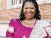 Nigerian Professor Jailed 70 Months For $1.4m Fraud In US Smiling woman in a purple dress with a patterned shawl, standing outside a brick building.