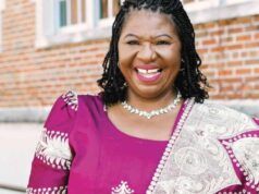 Nigerian Professor Jailed 70 Months For $1.4m Fraud In US Smiling woman in a purple dress with a patterned shawl, standing outside a brick building.