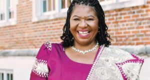 Nigerian Professor Jailed 70 Months For $1.4m Fraud In US Smiling woman in a purple dress with a patterned shawl, standing outside a brick building.