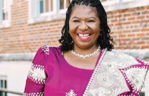 Nigerian Professor Jailed 70 Months For $1.4m Fraud In US Smiling woman in a purple dress with a patterned shawl, standing outside a brick building.