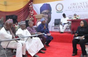OYRLEA Holds Farmers, Herders Dialogue On Enforcement On Open Grazing Officials seated on a red-carpeted stage at a government event, with a moderator speaking into a microphone on a podium in the background.