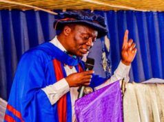 Ojo-Lanre Charges IIJ Matriculating Students To Embrace Development Journalism In The Age Of Phone Journalism Man in blue academic gown and cap delivering a speech at a podium, microphone in hand, raising one finger.