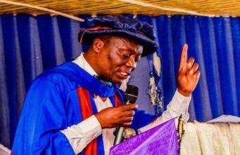 Ojo-Lanre Charges IIJ Matriculating Students to Embrace Development Journalism in the Age of Phone Journalism Man in blue academic gown and cap delivering a speech at a podium, microphone in hand, raising one finger.