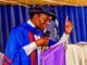 Ojo-Lanre Charges IIJ Matriculating Students To Embrace Development Journalism In The Age Of Phone Journalism Man in blue academic gown and cap delivering a speech at a podium, microphone in hand, raising one finger.