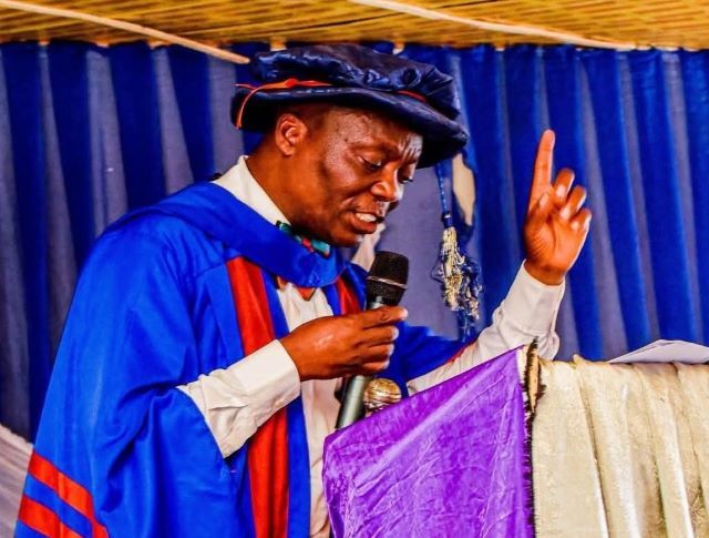 Ojo-Lanre IIJ Man in blue academic gown and cap delivering a speech at a podium, microphone in hand, raising one finger.