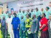 Oyo Trains Climate Change Desk Officers Across The State Group of diverse officials and attendees posing for a formal group photo indoors, with a banner and a line of people in teal uniforms visible in the background.