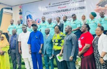 Oyo Trains Climate Change Desk Officers Across The State Group of diverse officials and attendees posing for a formal group photo indoors, with a banner and a line of people in teal uniforms visible in the background.