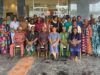 Oyo PHC Board Advances Family Planning Through SHINE Project Large diverse group of adults in colorful traditional clothing posing for a group photo outside a glass-front building; some seated in front and others standing behind them.