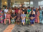 Oyo PHC Board Advances Family Planning Through SHINE Project Large diverse group of adults in colorful traditional clothing posing for a group photo outside a glass-front building; some seated in front and others standing behind them.