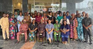Oyo PHC Board Advances Family Planning Through SHINE Project Large diverse group of adults in colorful traditional clothing posing for a group photo outside a glass-front building; some seated in front and others standing behind them.