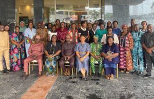 Oyo PHC Board Advances Family Planning Through SHINE Project Large diverse group of adults in colorful traditional clothing posing for a group photo outside a glass-front building; some seated in front and others standing behind them.